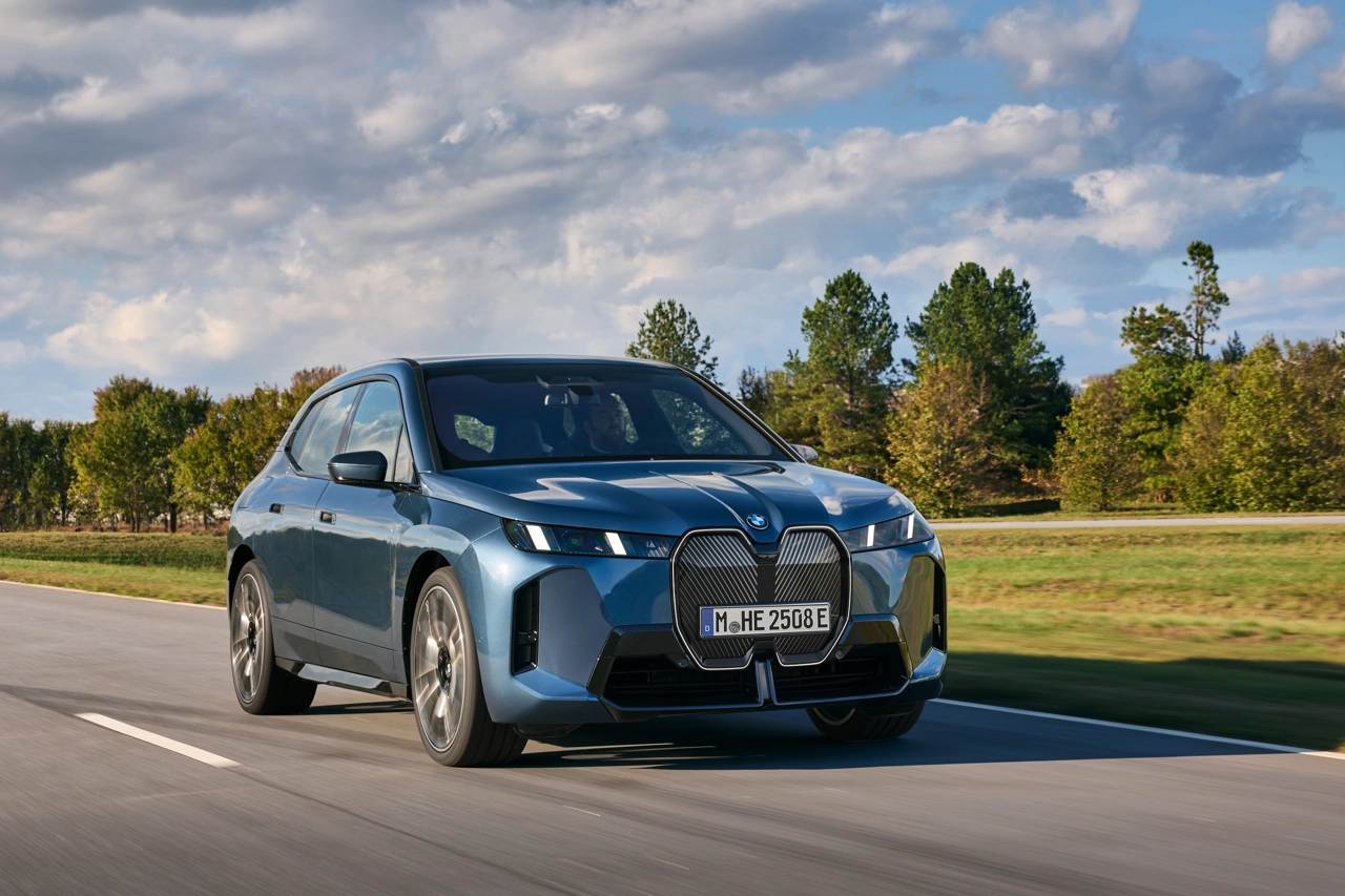 2026 BMW iX xDrive60 metallic blue in motion on a paved tree-lined road under a slightly cloudy sky.