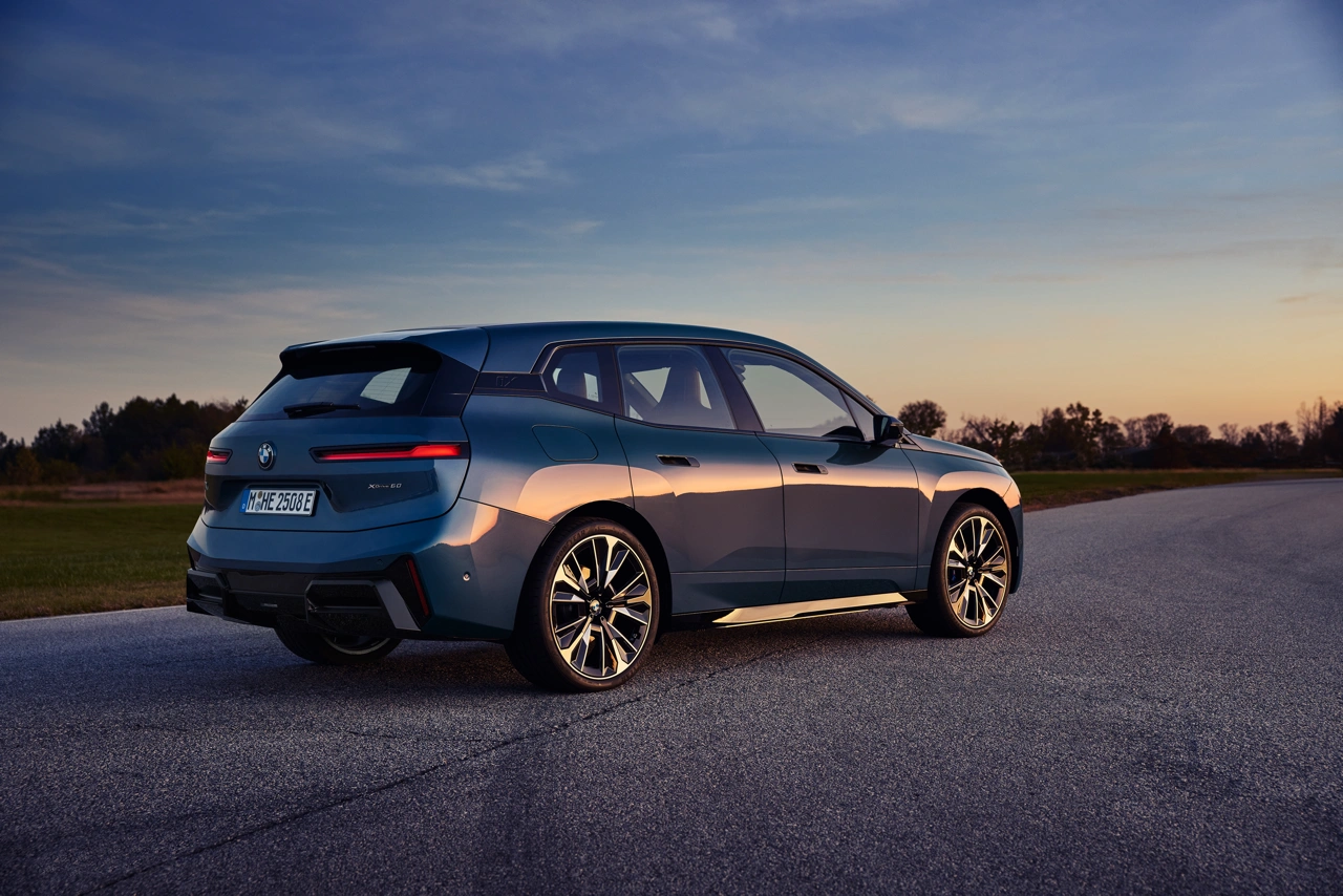 2026 BMW iX xDrive60 metallic blue rear three-quarter view on a country road at dusk with illuminated taillights.