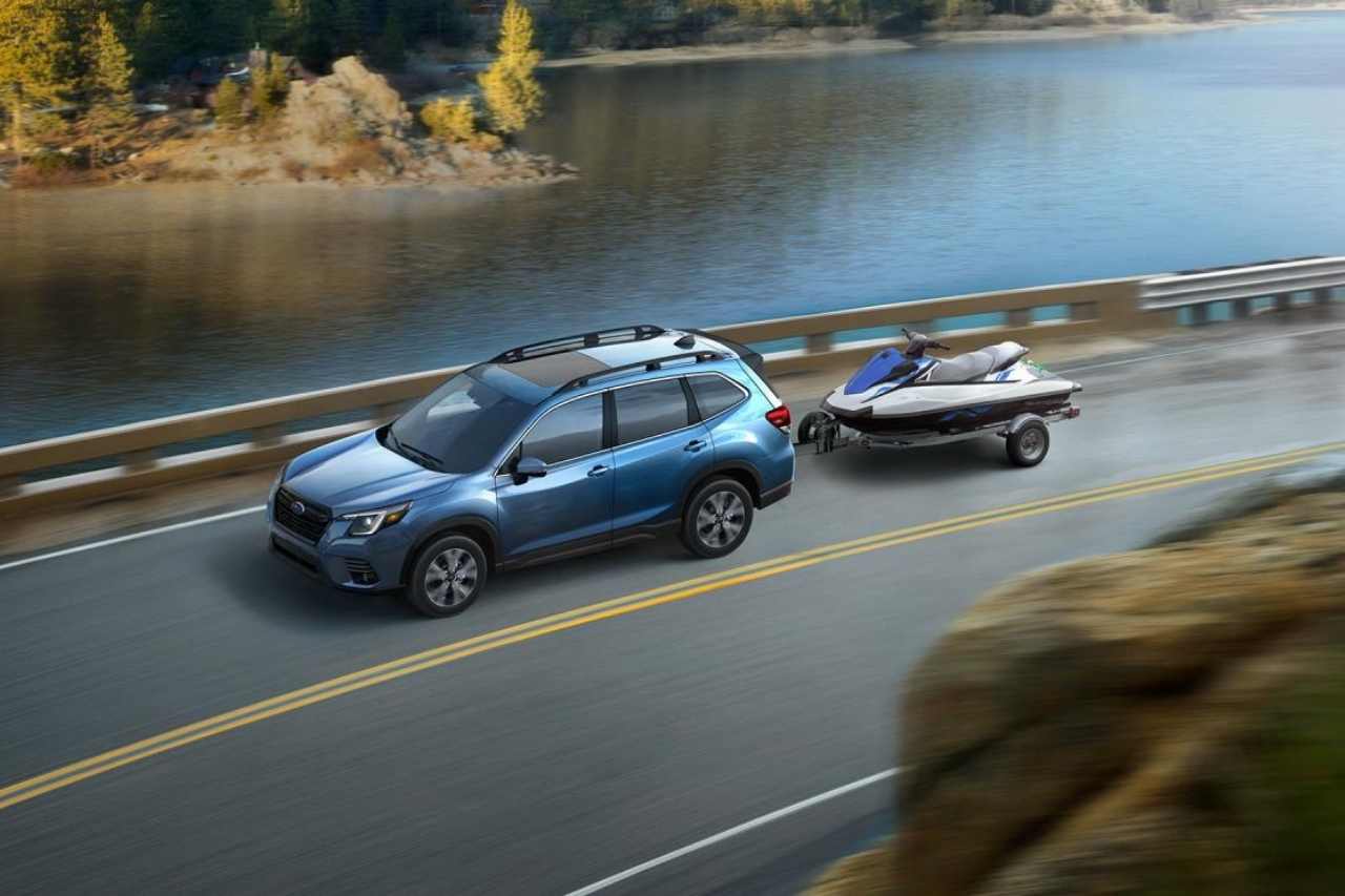 A blue Subaru SUV pulls a see-doo water vehicle along a road next to a lake.