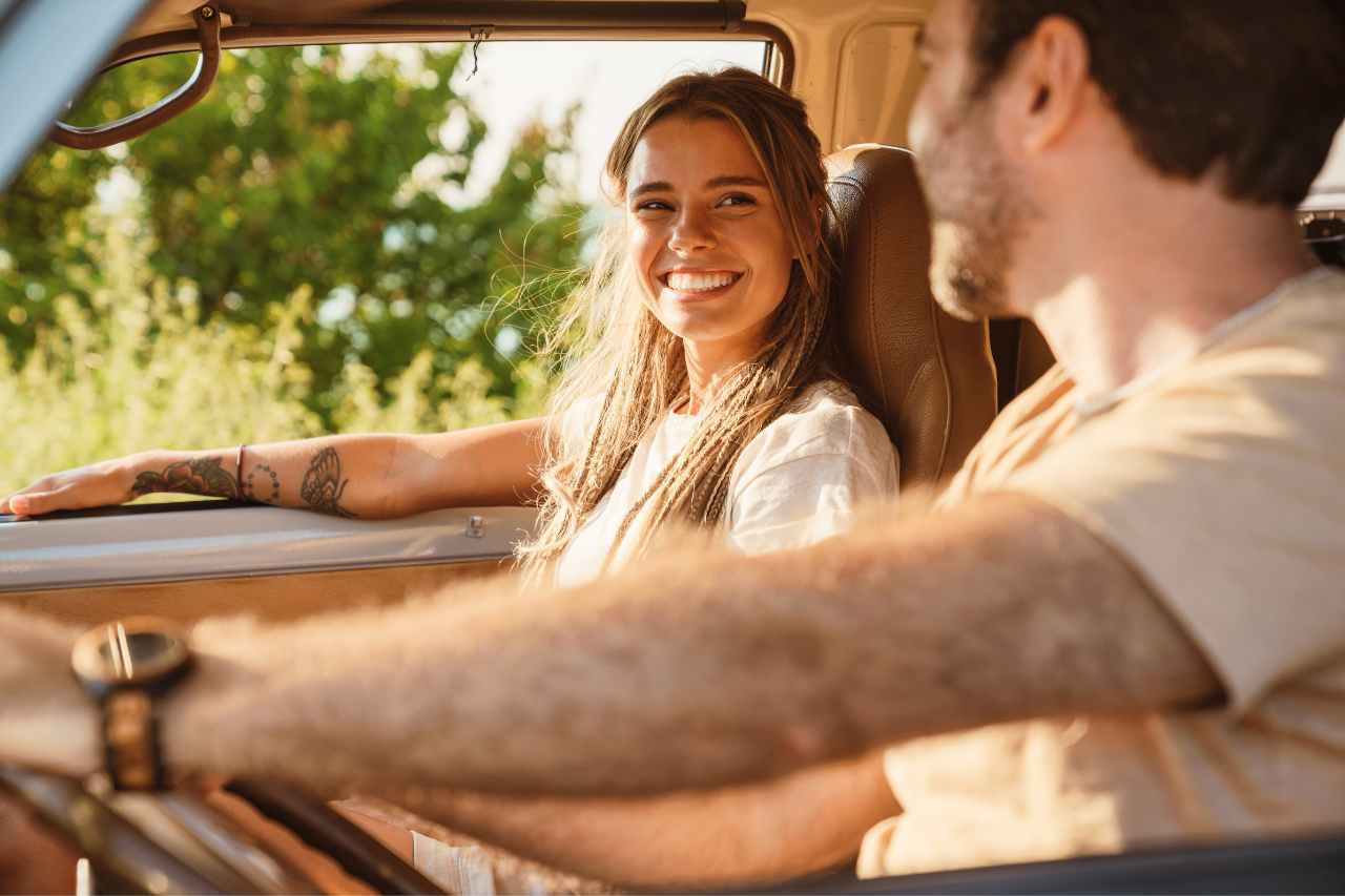 A smiling Caucasian man and woman drive down a sunny road with the windows down on a summer road trip.