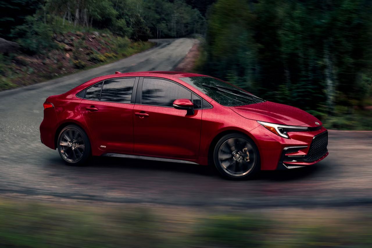 A red Toyota hybrid sedan drives down a rainy highway with a forest in the background.