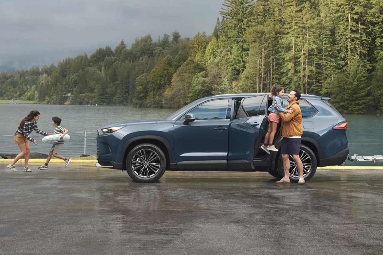 A blue-grey hybrid Grand Highlander SUV is parked next to a lake as a young family plays.