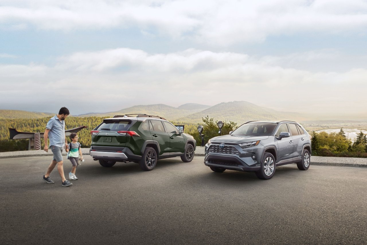 A man and his daughter walk past two Toyota RAV4s in a parking lot on top of a scenic hill. Green hills can be seen in the background.