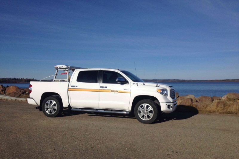 A white Toyota truck is parked on a road next to a large body of water on a sunny day.